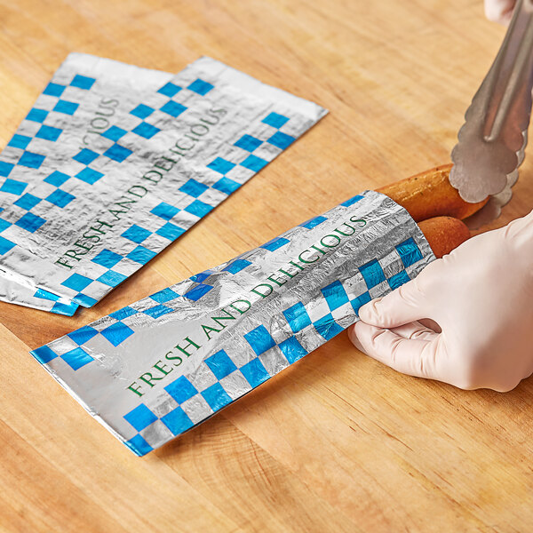 A person wearing gloves wrapping a hot dog in a Bagcraft foil wrapper with the words "Fresh and Delicious" and a honeycomb pattern.