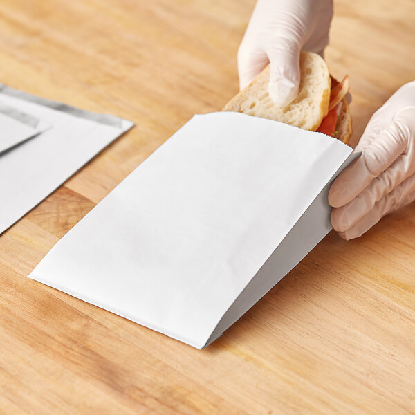 A person in gloves holding a white Bagcraft foil insulated sandwich bag with a sandwich inside.