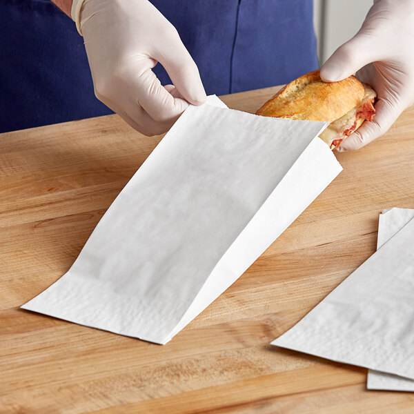 A person in gloves putting a sandwich in a white Bagcraft paper bag.