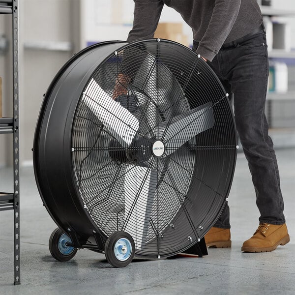 A man holding a Boltic 36" black portable drum fan with wheels.