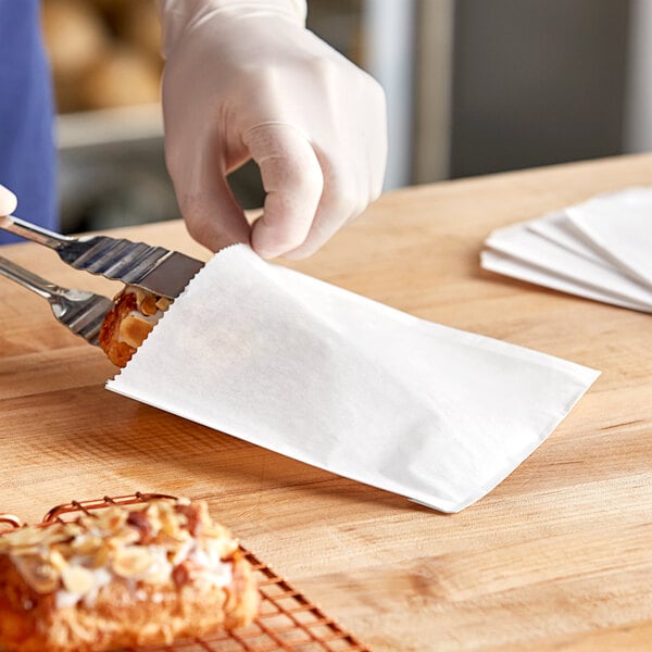 A gloved hand using tongs to place a pastry in a white Bagcraft sandwich bag.