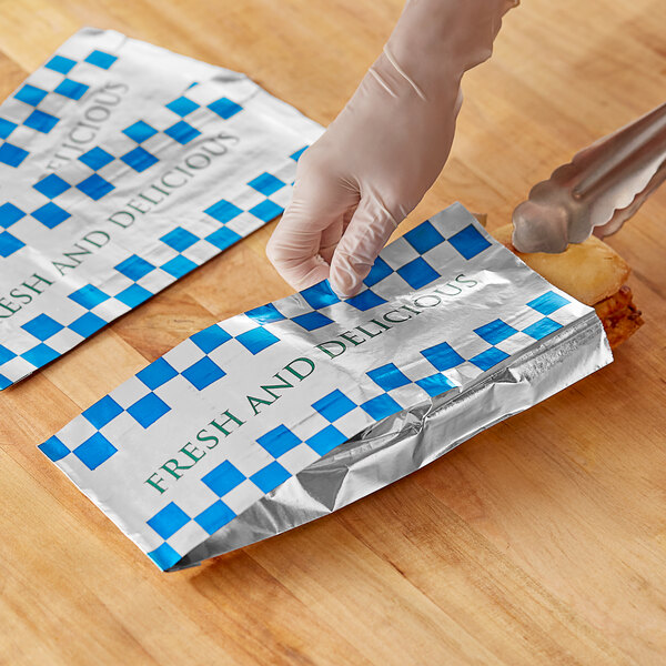 A person wearing gloves using a metal tong to put a sandwich in a Bagcraft foil meal bag.