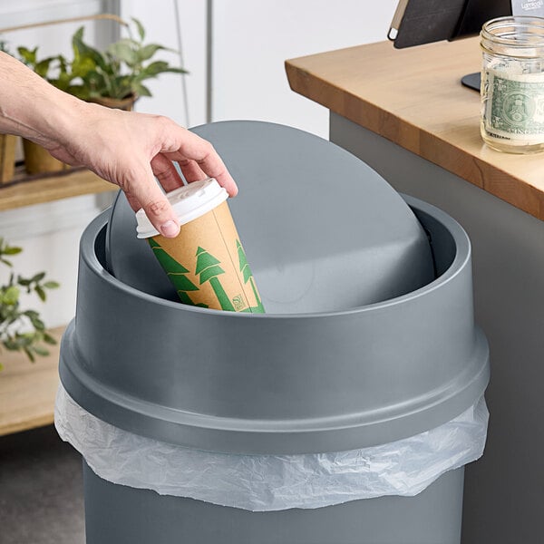 A hand putting a coffee cup into a Lavex swing-top wastebasket.