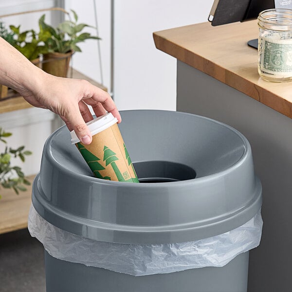 A hand pouring coffee from a paper cup into a Lavex round funnel wastebasket with a plant in the background.