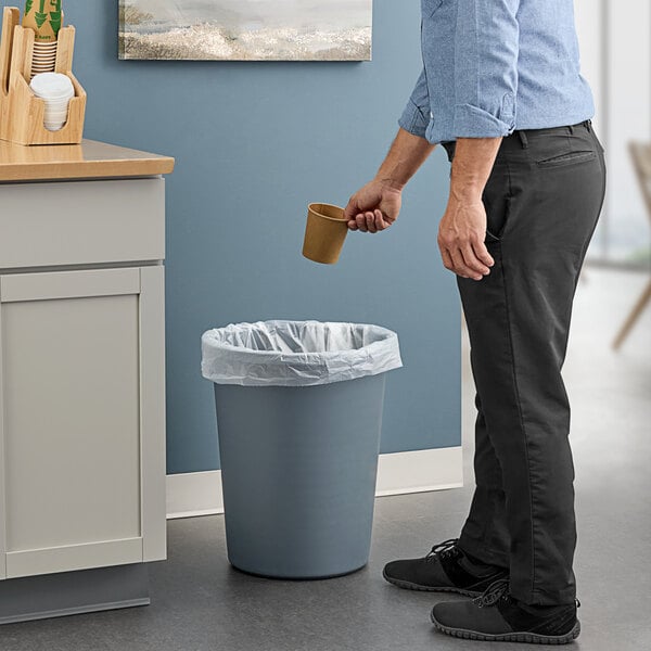 A man in a professional kitchen pouring coffee into a Lavex gray round wastebasket.