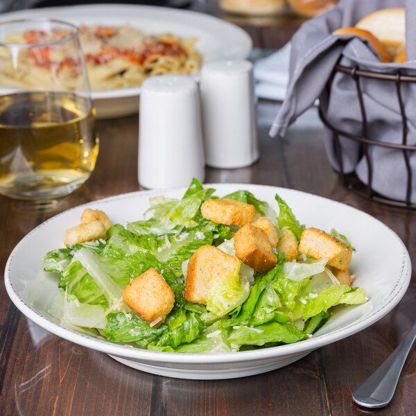 A round cream porcelain bowl filled with Caesar salad, featuring romaine lettuce, croutons, and grated cheese.