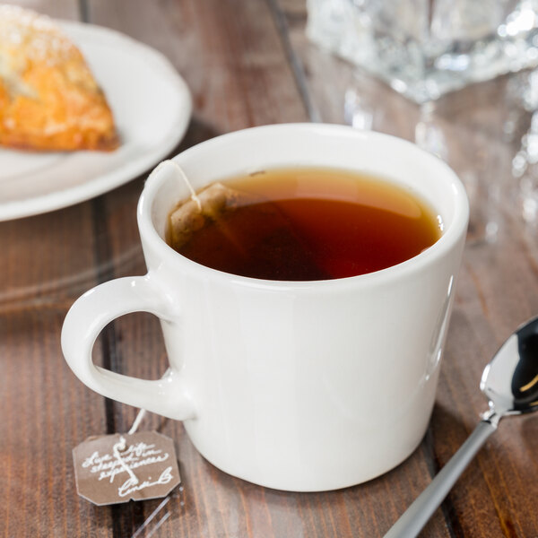 A Libbey Farmhouse porcelain cup with cream tea on a wooden table with a spoon and pastry.