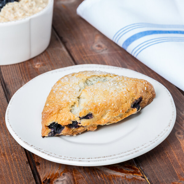 A blueberry scone on a Libbey Farmhouse round cream plate on a counter in a bakery display.