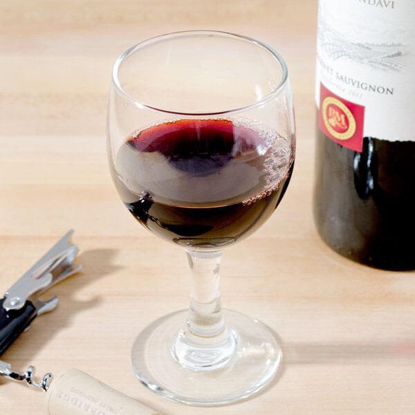 A Libbey wine glass filled with red wine next to a bottle of wine.