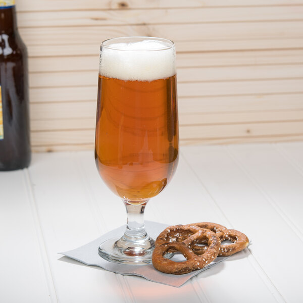 A Libbey stemmed pilsner glass of beer with pretzels on a table.