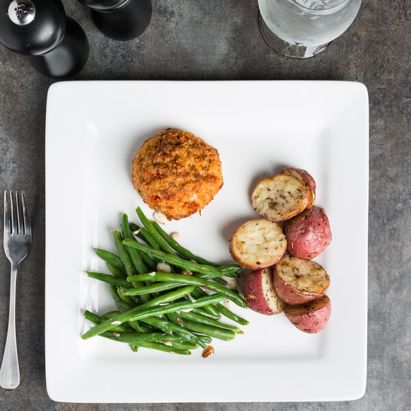 A white square porcelain plate with a serving of green beans, roasted potatoes, and a breaded patty.
