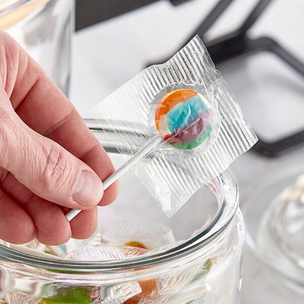 A hand holding a Clown Pops Tutti Frutti lollipop in a glass jar.