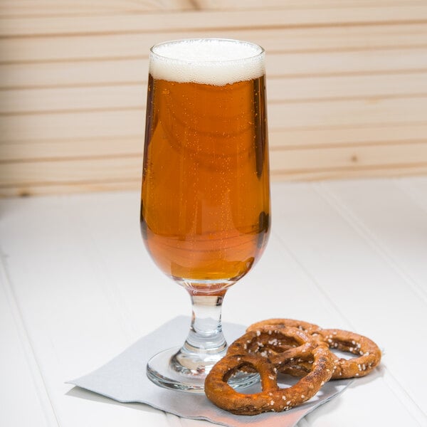 A Libbey stemmed pilsner glass of beer and pretzels on a table.