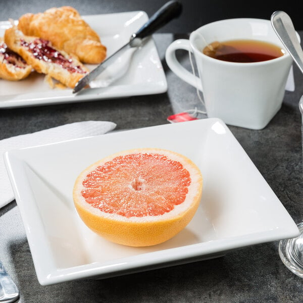 A Libbey wide rim square white porcelain bowl with half a grapefruit and a spoon on a table with a cup of tea.