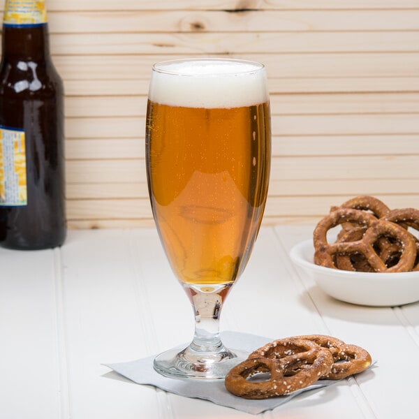 A Libbey stemmed pilsner glass filled with beer sitting on a table with a bowl of pretzels.