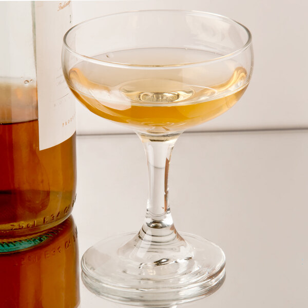 A Libbey Embassy coupe glass filled with liquid next to a bottle.