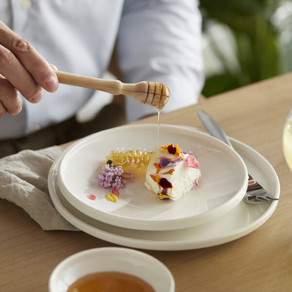 A person using a wooden honey dipper to drizzle honey over a piece of cake on a Libbey white porcelain plate.