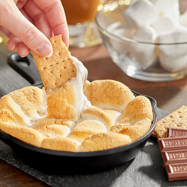 A person dipping a s'mores into a skillet of Campfire Large White Marshmallows.