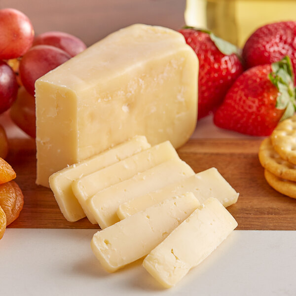 A wedge of Sartori BellaVitano cheese on a wood surface with strawberries and grapes.