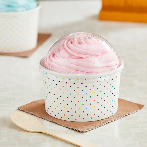 A white polka dot paper cup with a clear plastic lid filled with ice cream with a wooden spoon.