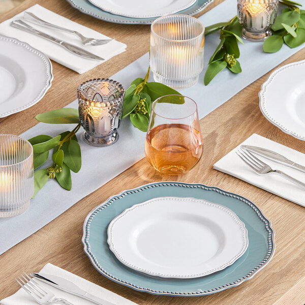 A table set with Visions white plastic plates and silverware on a white tablecloth with candles and greenery.