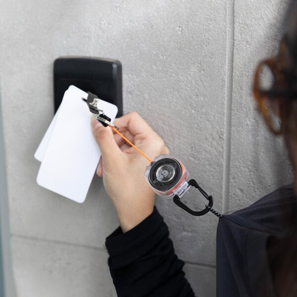 A person holding a KEY-BAK clear ID badge holder with a carabiner and orange Kevlar cord.