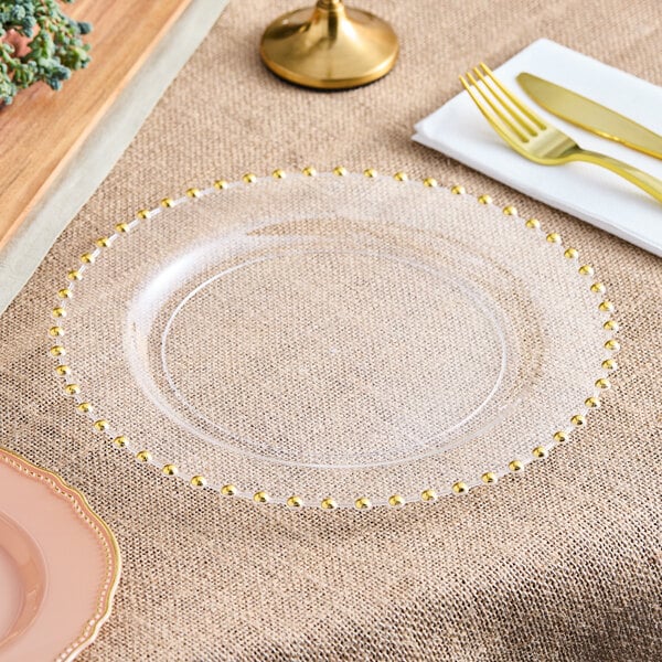 A Visions clear plastic plate with gold beaded trim on a table with a fork and knife.