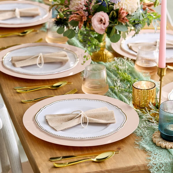 A wooden table is elegantly set with white plates featuring gold rims, gold cutlery, beige napkins tied with string, colored glassware, candles, and a floral centerpiece.