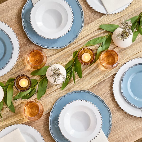 A table set with white Visions plastic plates with silver beaded rims and glasses of brown liquid on it.