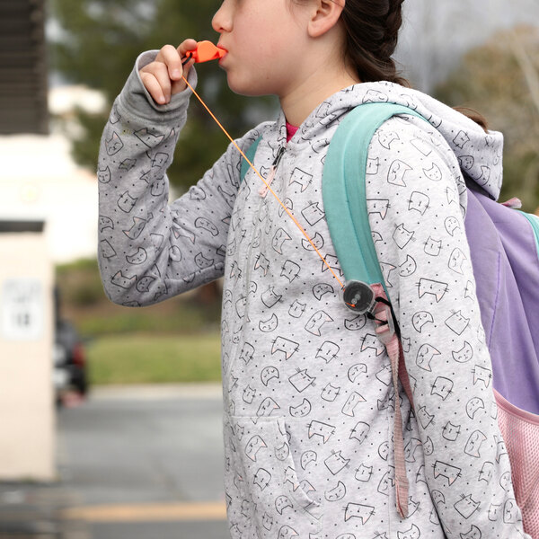 A girl wearing a backpack blowing an orange KEY-BAK whistle attached to her backpack.