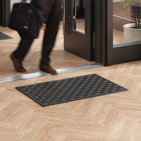 A person walking past a black rectangular Lavex entrance mat on a wood floor.