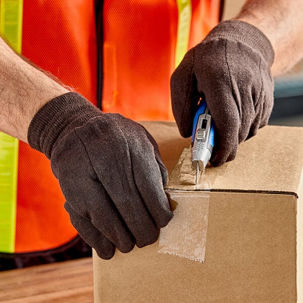 A person wearing Lavex brown jersey gloves using a box cutter to cut a box.