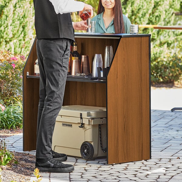 A man and woman standing next to a Lancaster Table and Seating foldaway bar with a walnut laminate finish.