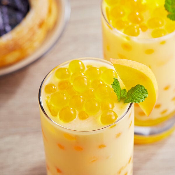 Two glasses of yellow bubble tea with orange Peach Popping Boba and mint leaves.