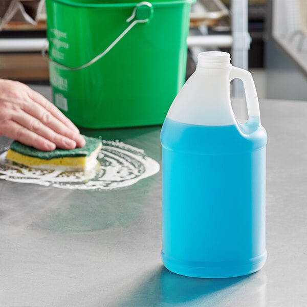 A person cleaning a counter in a school kitchen with a 0.5 gallon translucent blue jug of liquid.