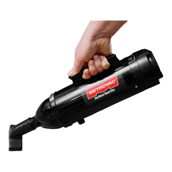 A hand holding a black MetroVac cordless handheld canister vacuum cleaner.