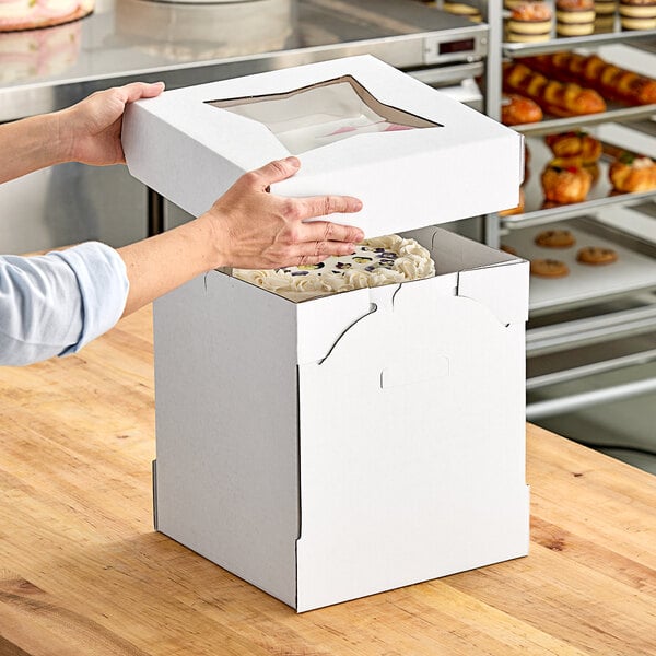 A hand opening a Southern Champion white cake box with a clear window to reveal a cake inside.