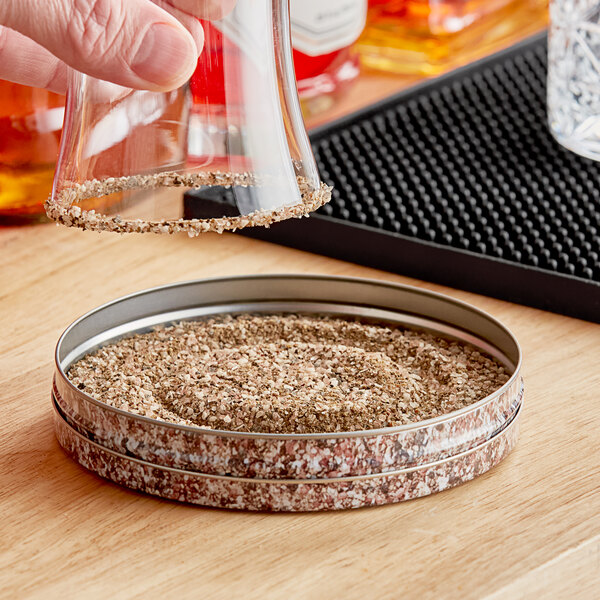 A hand pouring Demitri's Original Rim Shot Rimming Salt into a round container.