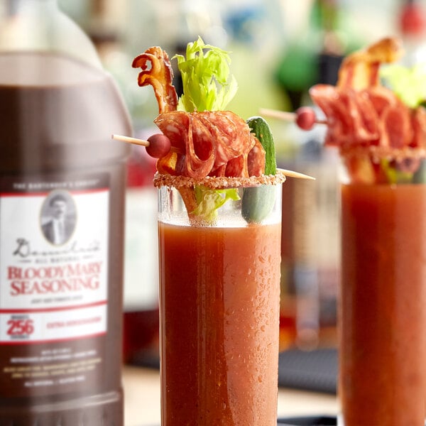 Two glasses of Demitri's Extra Horseradish Bloody Mary Concentrated Seasoning on a table with bacon and a bottle of liquor.