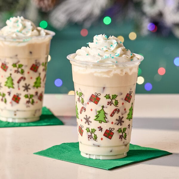 A 16 oz. clear plastic cold cup with holiday-themed decorations, filled with a creamy beverage and topped with whipped cream.