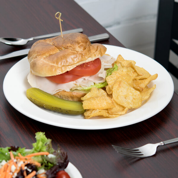 A Libbey white porcelain plate with a sandwich, pickles, and salad on a table.