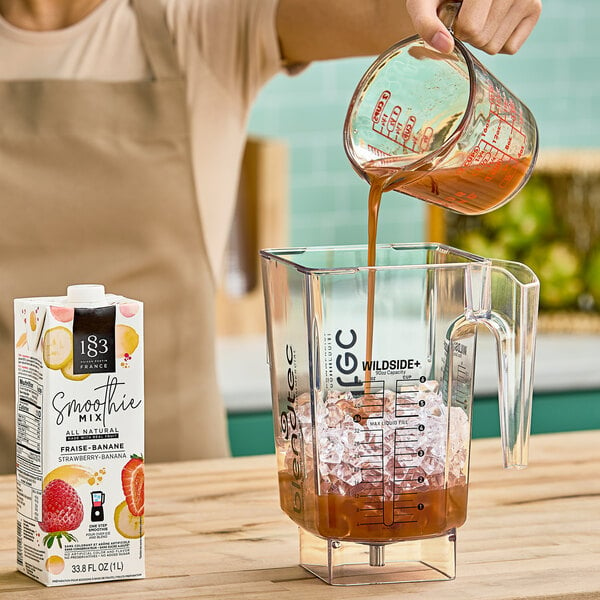 A woman pours 1883 Maison Routin Strawberry Banana smoothie mix into a blender.