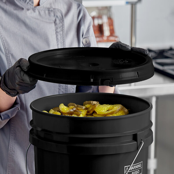 A woman in a chef's uniform holding a black BWAY pail cover with food inside.