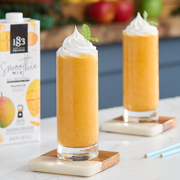 Two mango smoothies are sitting on a table next to a bottle of 1883 Maison Routin Mango Smoothie Mix.
