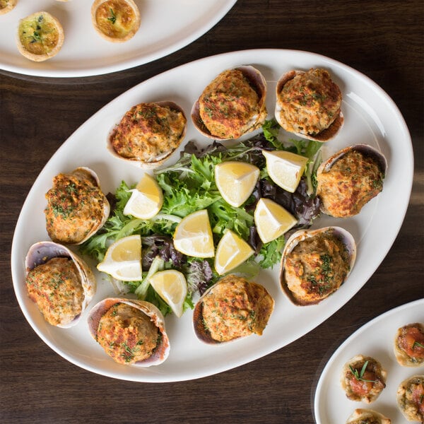 An oval white porcelain tray holding baked stuffed clams, garnished with lemon wedges and mixed greens.
