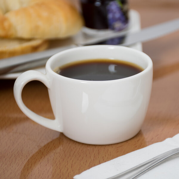 A Libbey white low espresso cup filled with coffee on a table next to a plate of bread.