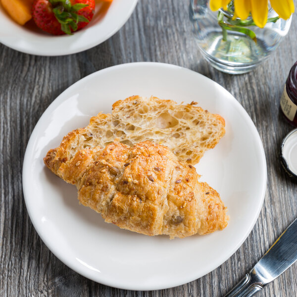 A Libbey round white coupe plate with a croissant and a knife on it next to a bowl of fruit.