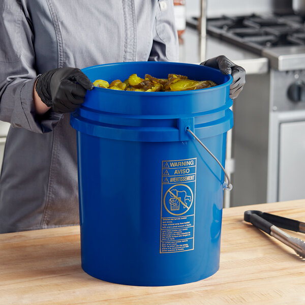 A woman in gloves holding a BWAY blue bucket full of food.