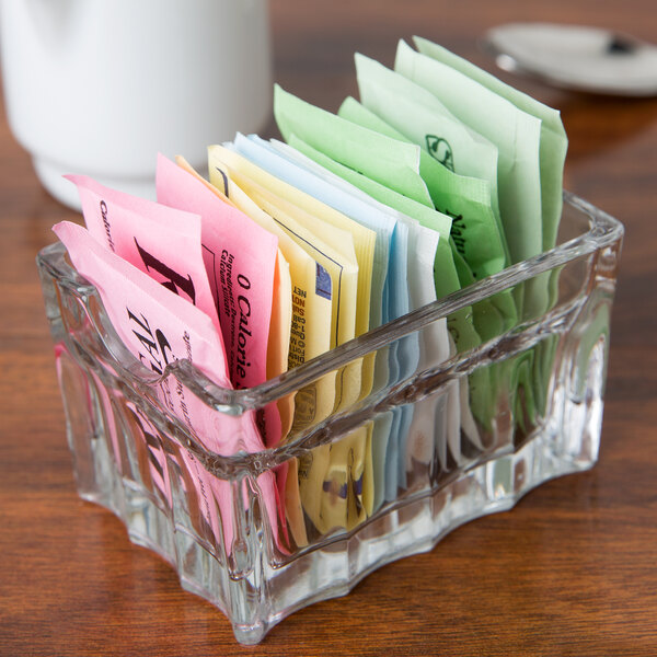 A clear Libbey glass container with different colored sugar packets in it.