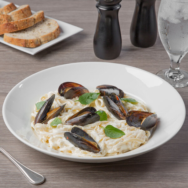 A Libbey white porcelain bowl filled with pasta, mussels, and basil leaves on a table.